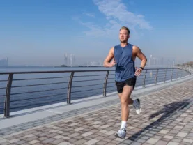 it man jogging along a waterfront path with a city skyline in the background, representing men's health and fitness.