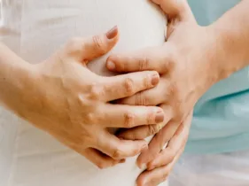 Close-up of a pregnant woman with her hands resting on her belly, joined by another pair of hands from behind, gently holding her belly.